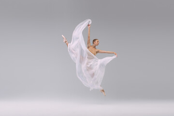Portrait of young beautiful ballerina dancing, jumping with transparent fabric isolated over grey studio background