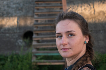 Looking straight into the camera, a thoughtfully smiling young woman of Slavic appearance with green blue eyes and braided brown hair in the setting sunlight against a wooden staircase and a concrete 