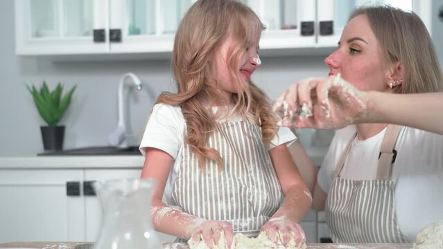 loving mom blows flour off her daughter dirty face while making dough at home in background of kitchen, close-up