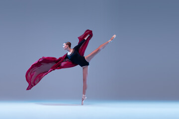 Portrait of young ballerina dancing with deep red fabric isolated over blue grey studio background. Standing on tiptoe
