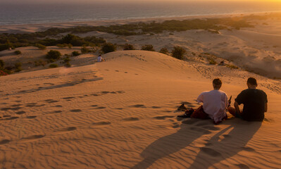 Young couple watching the sunset in the desert sand dunes, a woman and man sitting on sands at sunset