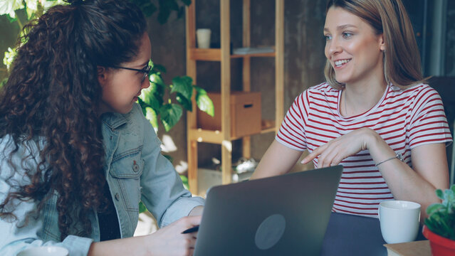 Young female enterpreneurs sitting together at table and talking emotionally. Women are wearing casual clothes and stylish accessories. Loft office in backfround.