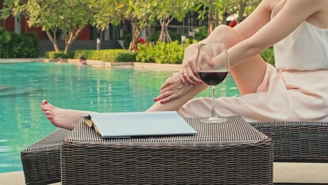Cropped shot of unrecognizable young woman in silk summer dress resting on sunbed by pool at luxury tropical resort with glass of red wine and good paper book