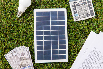energy saving, power and sustainability concept - close up of solar battery model, lightbulb, money with calculator and bills on green grass background