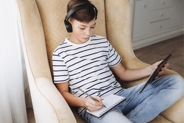 Teenage boy in wireless headphone using tablet studying online at home. Distance online education. Concept of e-learning, homeschool, virtual school.