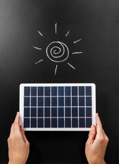 energy, power and sustainability concept - close up of hands holding solar battery model and sun on black chalkboard background