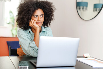 Bored tired african-american woman using laptop for remote work sitting in the living room, female student or freelancer feeling weary or lacks interest in current activity