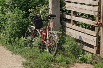 Fototapeta na wymiar one old red bicycle stands on the street in green grass and vegetation near a gray wooden fence