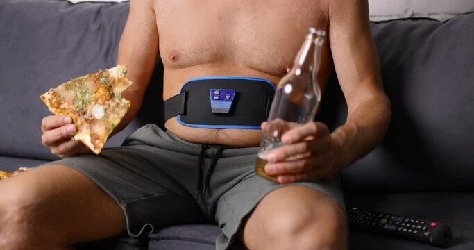 man sitting on couch eat pizza and drink beer while using electronic abdominal slimming belt. unhealthy lifestyle