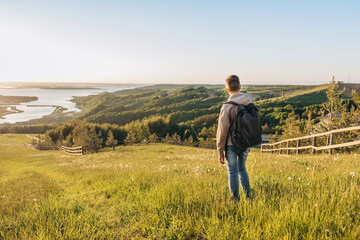 Naklejka na ściany i meble Tourist with backpack standing on top of hill in grass field and enjoying beautiful landscape view. Rear view of teenage boy hiker resting in nature. Active lifestyle. Concept of local travel