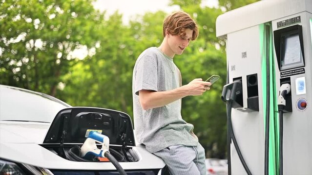 A young man using smartphone at a car charging station with charging electric car nearby. Chisinau, Moldova. Slow motion