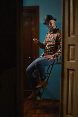 Handsome caucasian man in leather hat siting on ladder for repair and smoking cigarette.