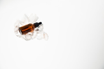 Fototapeta na wymiar An amber bottle with pipettte on a white feather on a white table with face serum product. Glass bottle with self care product. Top view