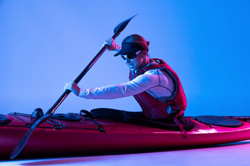 Water sport activities. Sportive woman in red canoe, kayak with a life vest and a paddle isolated on blue background in neon light. Concept of sport