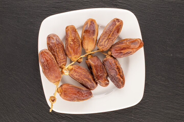 One branch with dry sweet dates on a white ceramic saucer on a slate stone, close-up, top view.