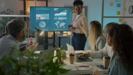 African American woman company manager presenting business ideas to diverse group of colleagues pointing at digital screen in office at night - Powered by Adobe