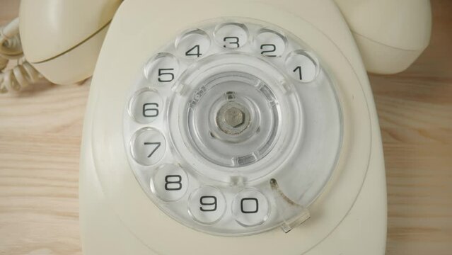 Old white rotary telephone with twisted cord on beige wooden table. Retro landline phone with rotary dialer and handset. Vintage analog plastic rotary telephone with dialing disk. Close up.