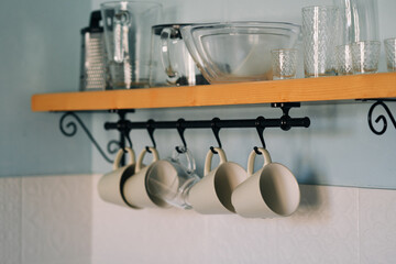 Kitchen open wooden shelf with dishes, glasses and mugs. Mugs are hanging from hooks. Cozy interior in a country house. Selective soft focus