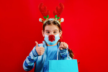 Child in wearing deer antlers and a medical mask with a nose t for Christmas with colorful shopping bag is isolated on a red background. Holidays concept. Holidays sales, shopping. Mock up bag in hand