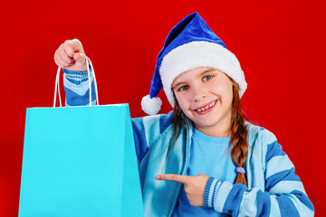 Cute child girl in a blue Santa's hat for Christmas with colorful shopping bag is isolated on a red background. Holidays concept. Holidays sales and shopping. Mockup bag in hand.