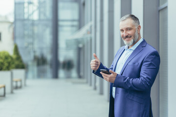 Portrait of successful senior gray haired banker, arab man looking at camera smiling and holding finger up, businessman outside office using smartphone