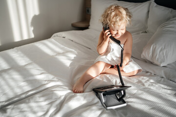 blonde child with curly blond hair rings the phone in a hotel room while sitting on a bed in a...