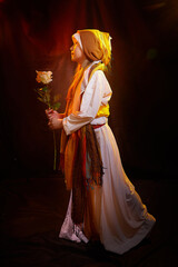 Little skinny girl in long white and yellow dress with scarf and flower. Young model posing for photo shoot in dark studio in stylized Arabic costume of Ancient East. Israel or Palestine teenager