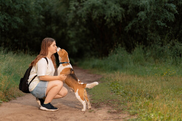 Teenage girl sitting on country road and hugging dog, side view. Beagle licking teenager face in rural