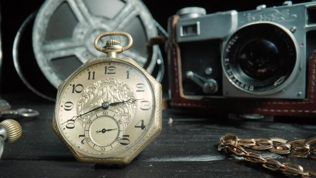 Antique gold and silver pocket watch, camera, gold chain and vintage film reel on black background. Retro pocket watch with ticking second hand. Vintage objects are laid out on dark table. Close up.