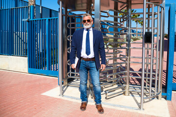 Mature, grey-haired, bearded professor with sunglasses stands at the entrance to the university with his laptop in his right hand. Concept of education and teaching. Teachers and retirement.