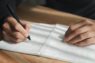 Close up of young man in casual cloth hands writing down on the notepad, notebook using ballpoint pen on the table.