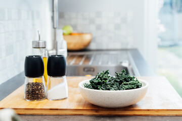 Teared curly green kale leaves in the plate, spice, salt and olive oil bottle on the kitchen table. Ingredients for kale chips or healthy salad meal. Healthy eating, dieting lifestyle. Selective focus
