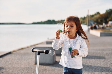 Playing with bubbles. Cute little girl with scooter is outdoors in summertime