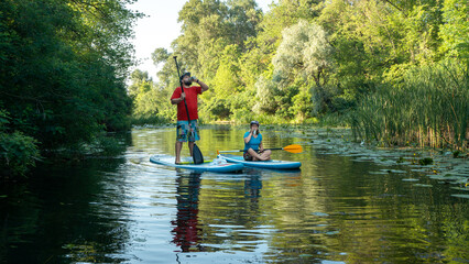 Fototapeta na wymiar A man with a beard and a woman stand on paddle boards on the river and drink. Summer time