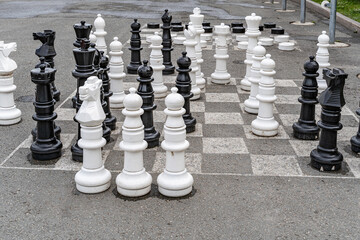 A chessboard on asphalt with white and black chess pieces for playing and competing in a chess battle on the square in front of the City Administration building near the park and a bench for fans.