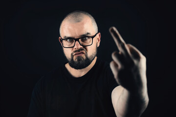 Portrait shot of bald bearded caucasian male with glasses showing middle finger on black background 