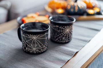Cozy halloween plans at home. Hot tea drink in black mugs with spider net pattern, cookies and and sweets on the plate, pumpkin decor on the coffee table. Festive autumn, fall mood home decor.