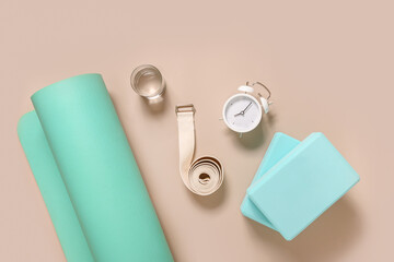 Green yoga mat, two blocks, water, clock and white belt on beige background with copy space. Top...