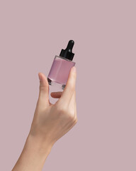 Woman hand holding facial essential oil or serum packaging on pink background. Beauty cosmetic...