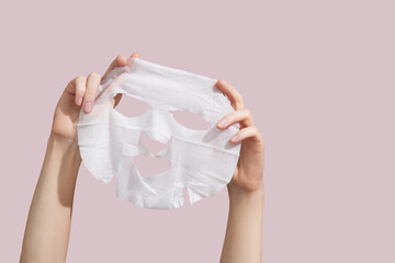 Female hands holding sheet of white mask on pink background. - obrazy, fototapety, plakaty