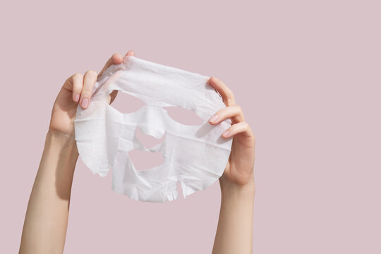 Female hands holding sheet of white mask on pink background.