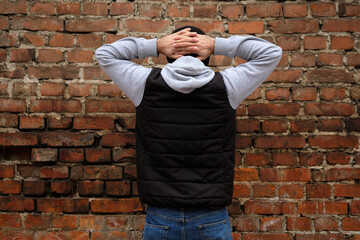 The guy stands facing a brick wall with his hands clasped behind his head. A guy in jeans and a...