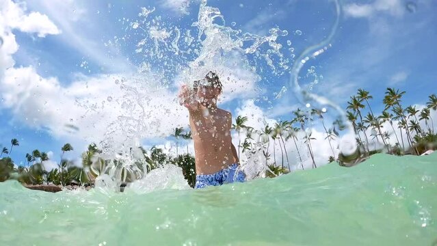 Half underwater shot of a young boy splashing clear sea water at the beautiful beach with palm trees