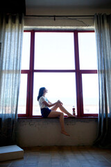 A girl in a white T-shirt and shorts is sitting at the window.