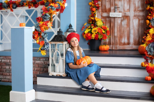 Little happy girl holding pumpkin and smiling at camera while sitting on house porch and waiting to trick or treat on Halloween. Child is sitting next to pumpkins near door of house. Autumn home decor