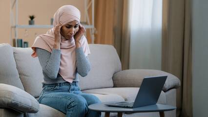 Young muslim woman in pink hijab sitting on sofa working using laptop having headache migraine sad female freelancer feel severe pain tired stressed high pressure overtime work health troubles concept