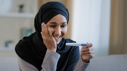 Portrait joyful surprised girl in hijab holding pregnancy test sit in living room excited with...