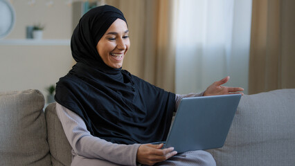 Friendly arabian muslim woman girl in hijab sit on cozy sofa talk video call with friend greeting at computer webcam take interview distance virtual communication in social networks use wi-fi at home