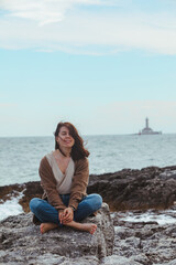 Fototapeta na wymiar woman sitting by rocky sea beach in wet jeans lighthouse on background