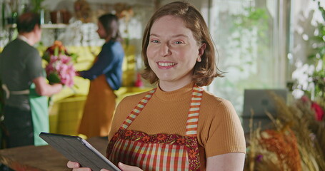 Portrait of a young female employee holding tablet smiling at camera standing inside small business flower shop store. Staff checking online orders looking at camera smiling